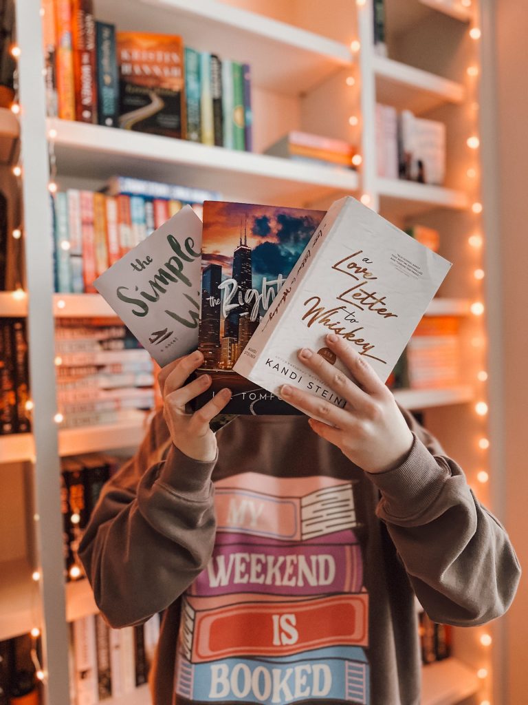 In front of her bookcase, illuminated with twinkle lights, Kailey, dressed in a brown bookish sweatshirt, holds up three romance titles. The only title completely visible is A Love Letter To Whiskey by Kandi Stein.