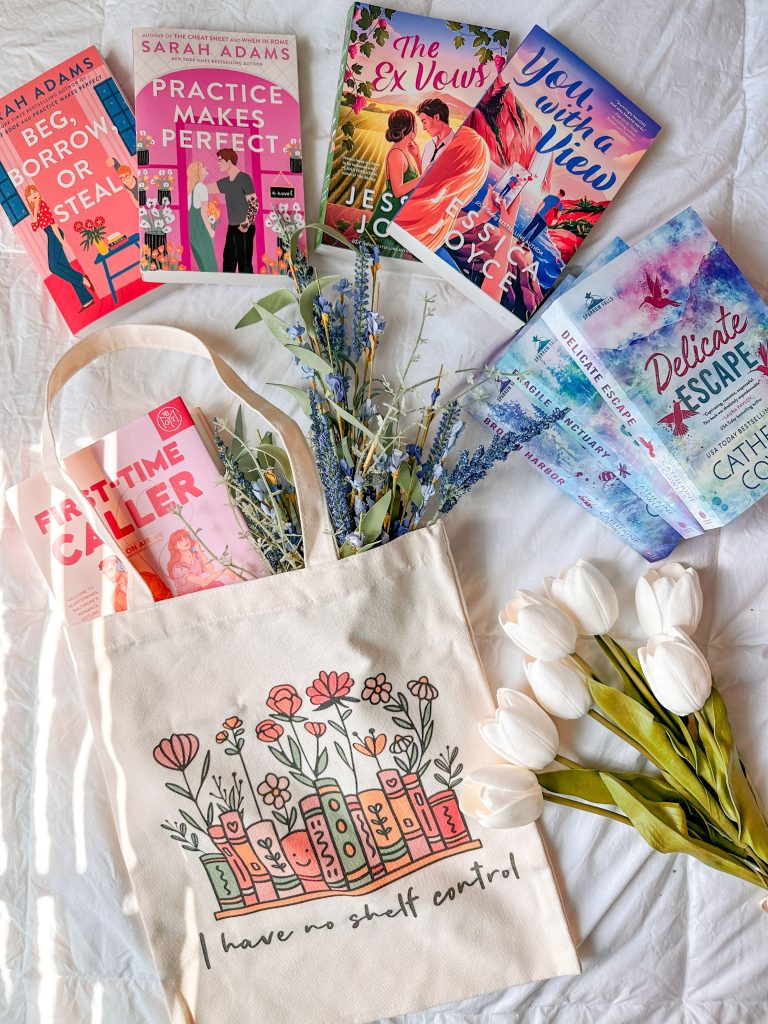 On top of a white sheet, a tote bag decorated with book bindings and flowers has a bouquet of blue flowers and the romance novel First Time Caller spilling out of it. Around the tote are several other romance novels, the author Jessica Joyce and the novel Delicate Escape are visible, and a bouquet of white tulips are in the bottom right corner.