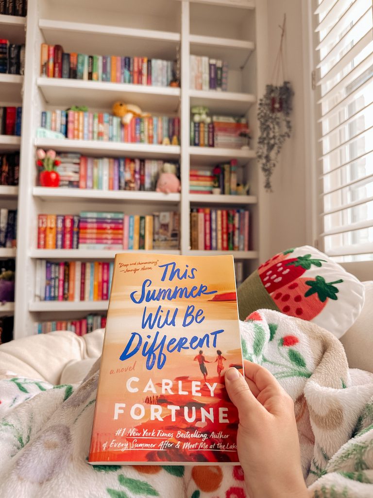 Laying in bed, Kailey's hand holds up the romance book cover This Summer Will Be Different by Carley Fortune as she snuggles under her cozy white blanket, decorated with strawberries. In the background, a strawberry pillow and Kailey's colorful bookshelf can be seen.