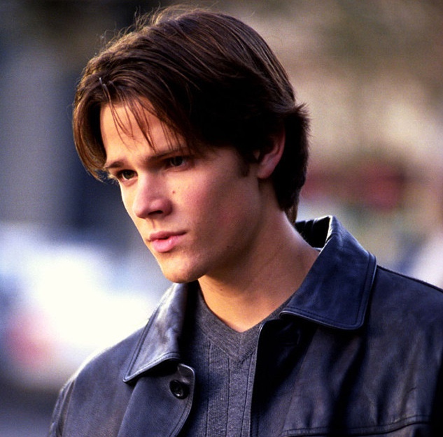 A still from Gilmore Girls. A young man with brown hair and pale skin wears a gray sweater and a black leather jacket.