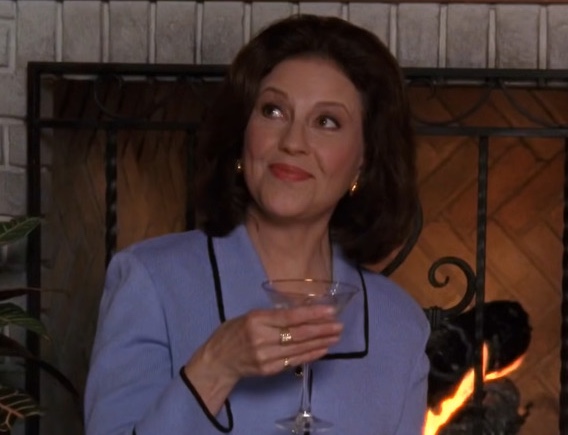 A still from Gilmore Girls. An older woman with dark brown and a powder blue suit holds a martini glass and sits in front of a gray stone fireplace.