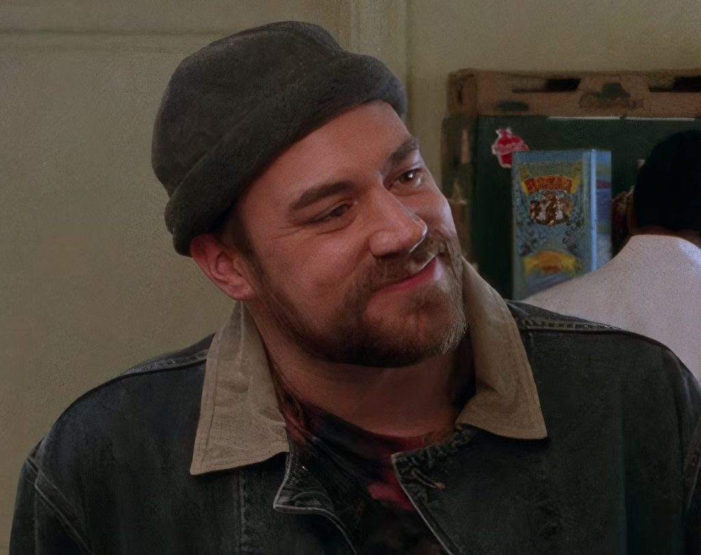 A still from Gilmore Girls. A man with pale skin and a brown beard wears a dark gray rolled bean and a dark denim jacket.