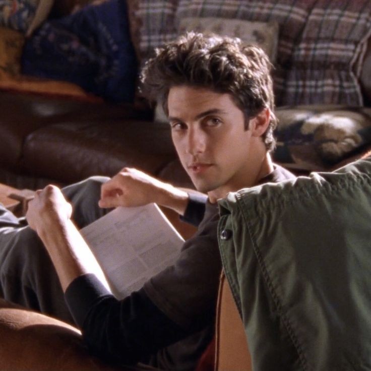 A still from Gilmore Girls. A young man with black wavy hair sits on a leather armchair and looks over his shoulder. In his lap is an open book. Behind him is a brown leather couch covered in multicolored blankets.