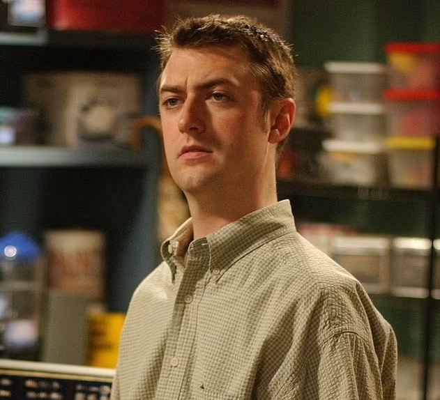 A still from Gilmore Girls. A man with pale skin and brown hair wears a green gingham shirt looks off in the distance with a furrowed brow. Behind him are supply shelves.