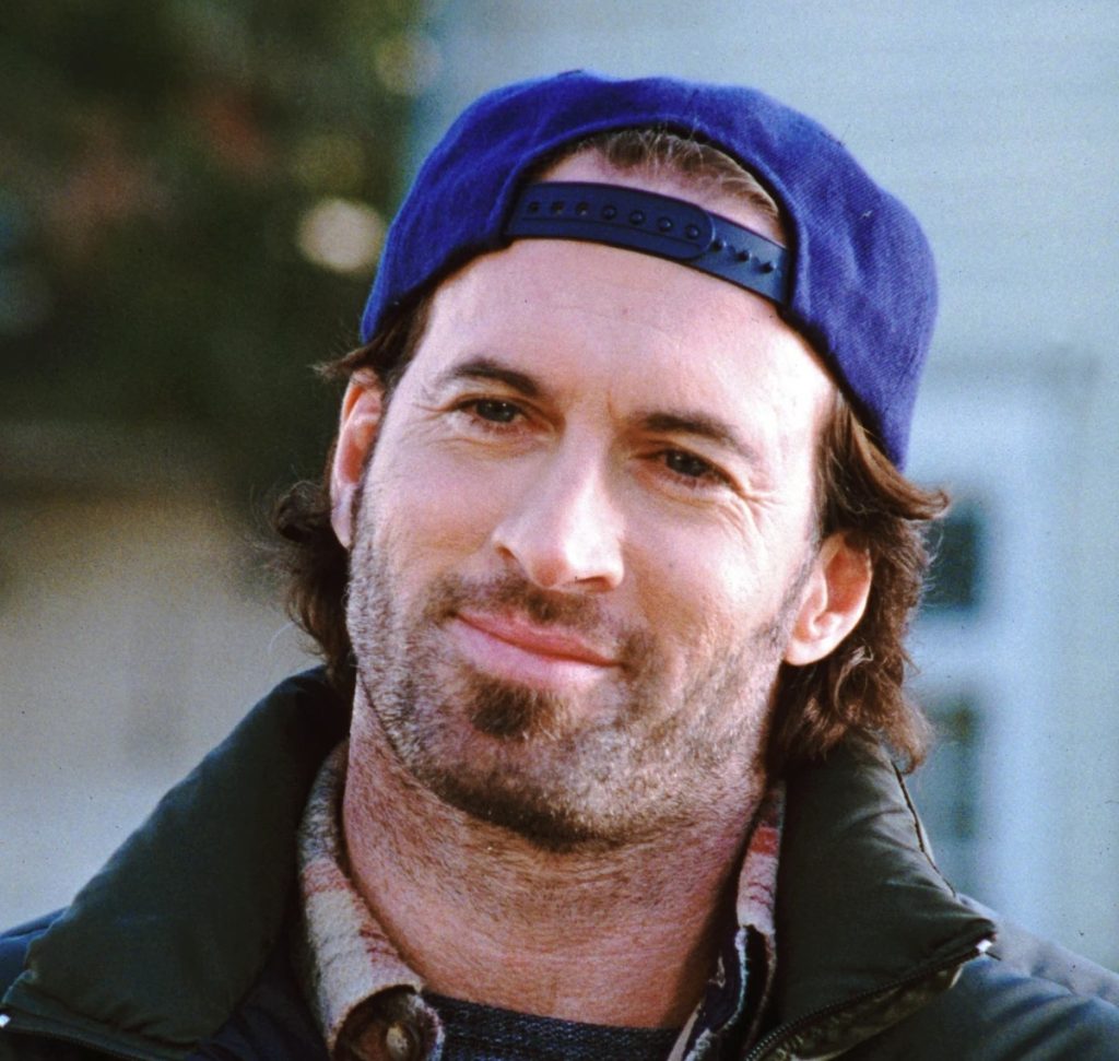 A still from Gilmore Girls. A man with brown hair and stubble wears a backwards blue baseball cap and smiles.