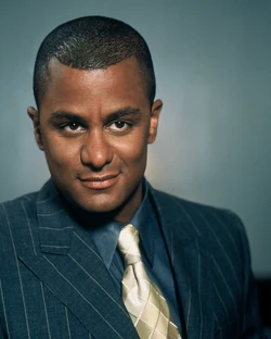 A promotional still from Gilmore Girls. A man with brown skin and a shaved head wears a dark pinstripe suit, a blue shirt, and a gold tie. He smolders in front of a gray-blue background.