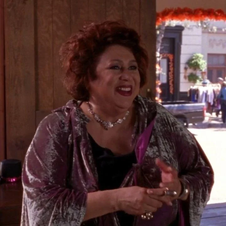 A still from Gilmore Girls. An older woman with reddish brown curly hair wears a purple velvety garment and smiles.