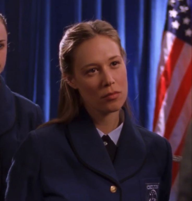 A still from Gilmore Girls. A young woman with golden blonde hair and pale skin wears a navy blue private school uniform and scowls. Behind her are blue curtains and an American flag.
