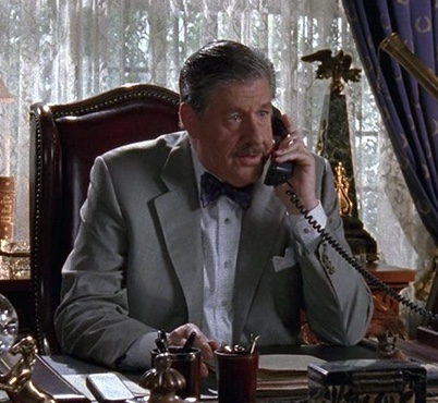A still from Gilmore Girls. An older man with gray hair and mustache wears a gray suit and dark bowtie. He sits in a burgundy leather chair and talks on a black desk phone. Around him is an opulently decorated desk and white lace window curtains.