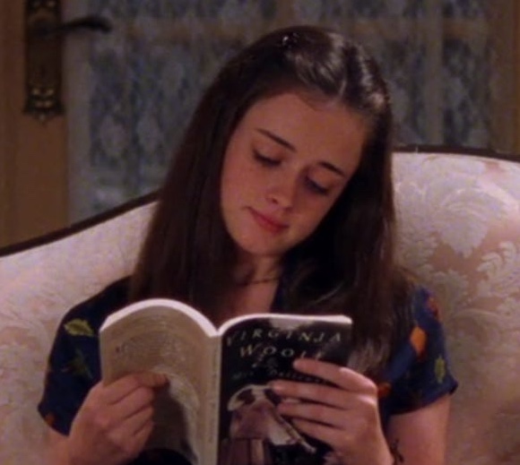 A still from Gilmore Girls. A young girl with dark brown hair wears a blue printed dress and sits on a pink antique couch. She reads a copy of Mrs. Dalloway by Virginia Woolf.