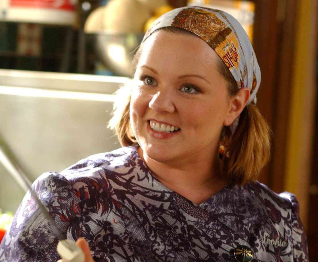 A still from Gilmore Girls. A woman with pale skin and golden blonde pigtails wears a pale blue bandana and a purple and red printed chef's coat. She gestures with a knife in her hand and smiles.