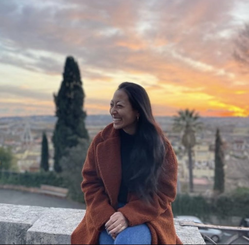 Natalie Naihuei Hsieh, an East Asian woman with long dark hair, an orange coat, and jeans, sits on a concrete wall, smiling with her face turned three-quarters to the viewer's left. A city landscape is visible behind her, dotted by tall trees and lit by a setting sun.