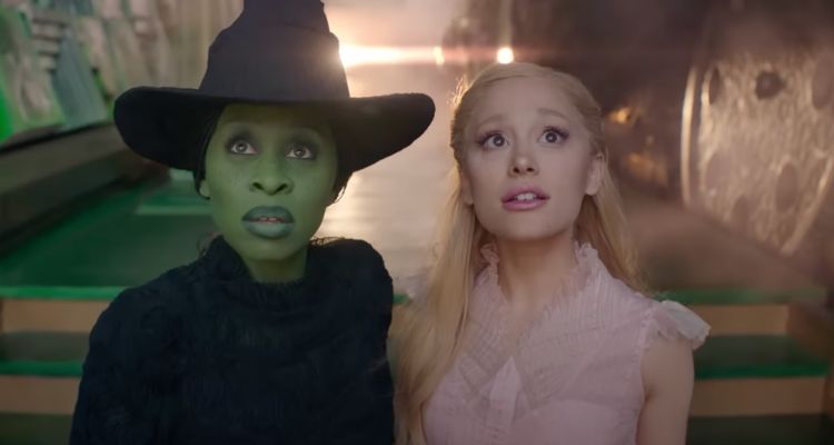 Still from Wicked (2024), depicting Elphaba (Cynthia Erivo) in a black outfit and black witch hat and Glinda (Ariana Grande) in a pink outfit. They are looking up at something above the camera, and the background is out of focus, but depicts green stairs and a bright light from behind them.