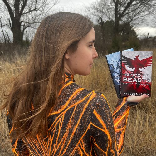 Rebecca Lemke's back profile, with her face slightly turned towards the camera. She has brunette hair, pale skin, and a painted black and yellow blazer. She is holding up the Blood of Beasts and Head Under Water books, and is in an outdoor setting.