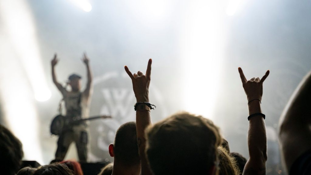 A crowd member at a rock and roll concert holds up their hands with the sign for rock and roll while a man with a guitar around his torso holds up the same hand signs on a stage.