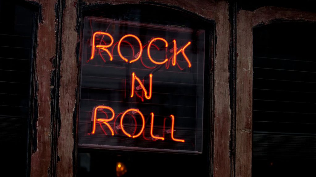 A red neon rock n roll sign hanging on a weathered wood wall.