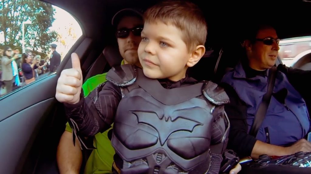 Still from Batkid Begins, depicting a young boy wearing a Batman costume in a car looking out of a window with a thumbs up. He is sitting on a man's lap, and another man is beside them. Outside the window, there is a large group of people.