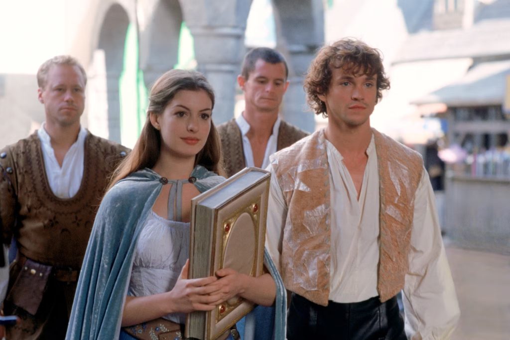 Still from Ella Enchanted, depicting Ella (Anne Hathaway) in a light blue dress with a velvet blue vintage cape and Prince Charmont (Hugh Dancy) in a beige vintage long-sleeve and pale orange vest standing together. Behind them are two men in brown vests.