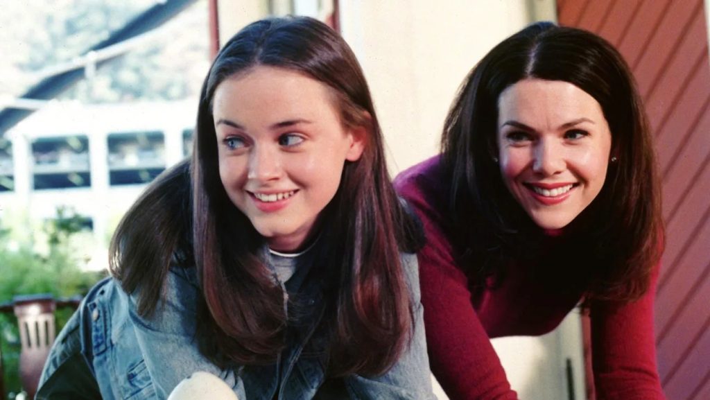 Still from Gilmore Girls depicting Rory Gilmore (Alexis Bledel) in a denim jacket leaning on something and smiling at something to her right. Next to her is Lorelai Gilmore (Lauren Graham) in a red long-sleeve top also looking to her right and smiling.