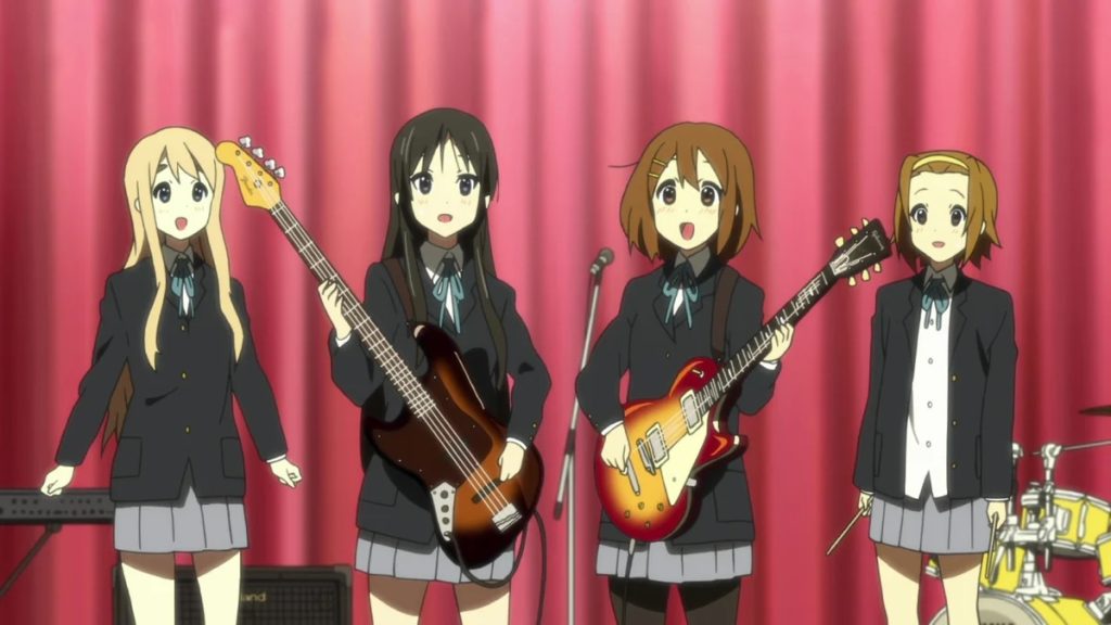 Still from K-On!, depicting four animated girls. Two are holding guitars and are in front of a mic stand, while the other two are standing in front of a keyboard and drums. They are all wearing black blazers, white blouses, blue bow ties, and grey pleated skirts.