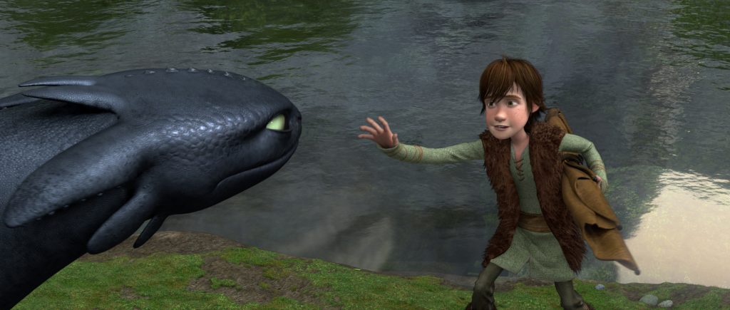 Still from How to Train Your Dragon (2010), depicting Hiccup, a pale, brown-haired boy in a brown vest and green outfit, sticking his arm out to Toothless, a black dragon with green eyes. The background depicts grass and a lake.