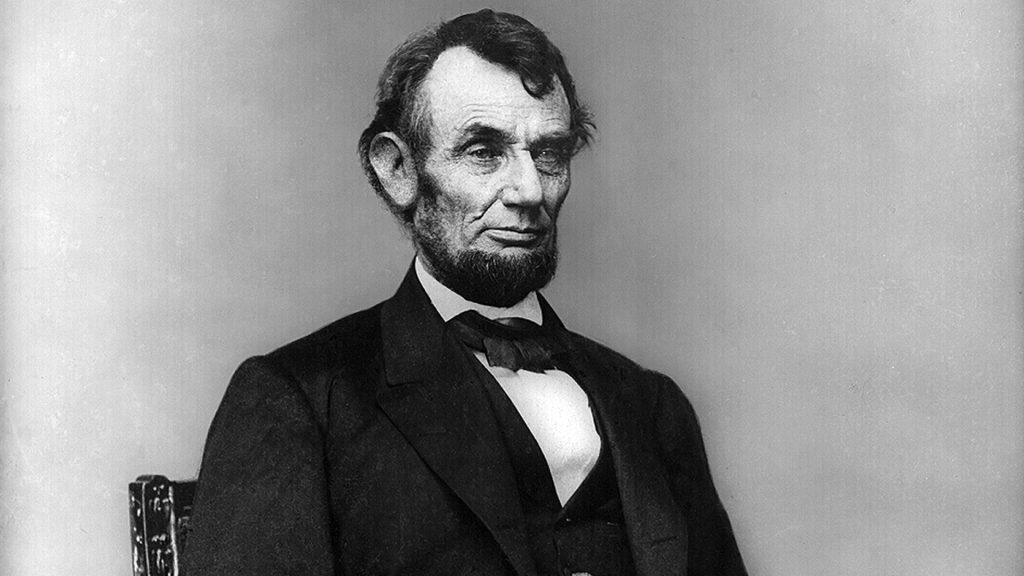 Portrait of Abraham Lincoln sitting in a chair, wearing a black suit, and looking off to the right with a blank facial expression.