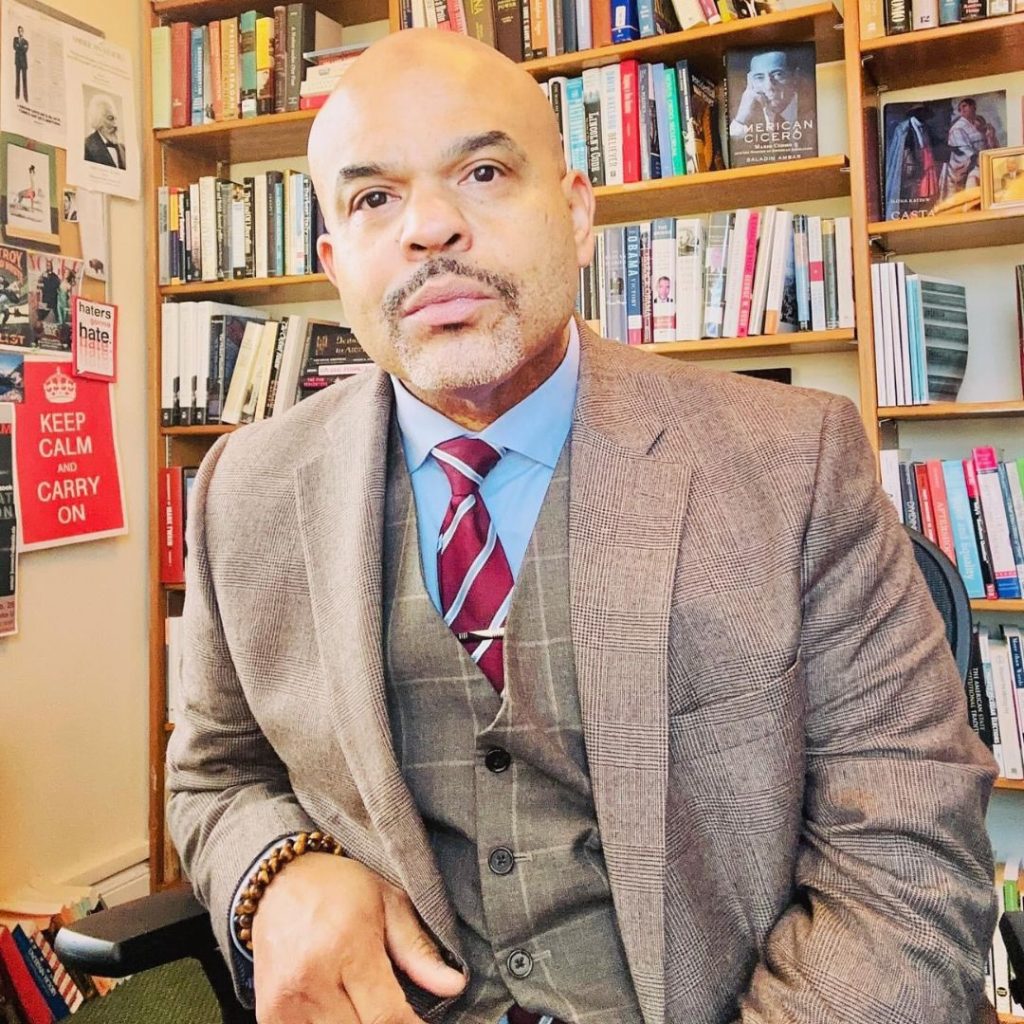 Saladin Ambar sitting in a chair in front of a bookshelf filled with books. He is bald with tan skin, short gray facial hair, and brown eyes. He wears a gray suit with a blue undershirt and white and red striped tie. His facial expression is neutral.