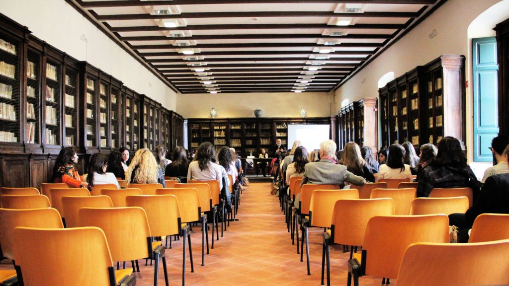 A long room with bookshelves lining the walls of the front half, framing a desk and projector screen at the head of the room. Lines of chairs extend past the camera's point of view with people sat in most of the chairs.
