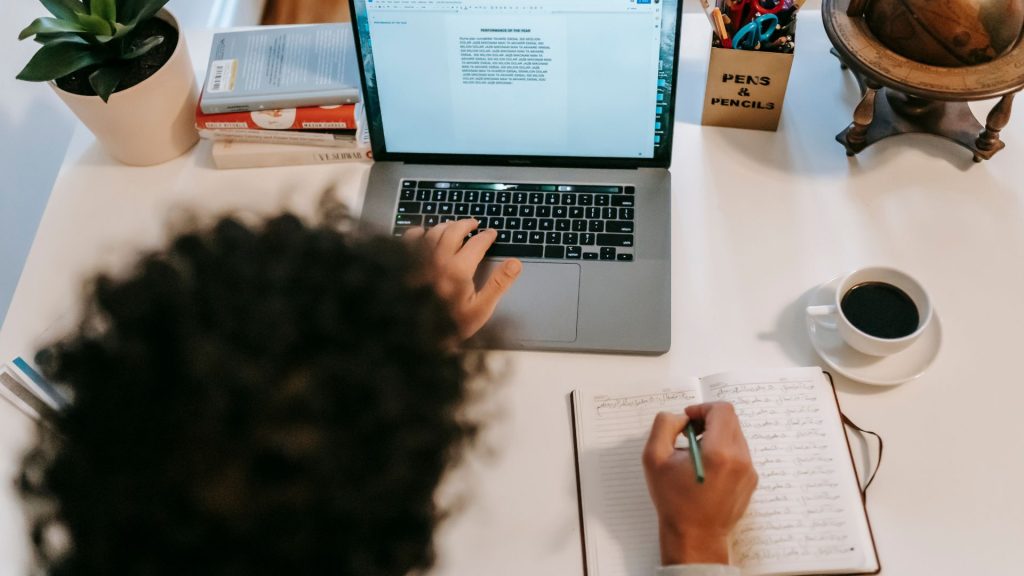 A person at their desk with their laptop, a stack of books, a caddy for writing utensils, a notebook, and a cup of coffee. On their computer, a word document is open as they're typing and taking notes in their notebook.