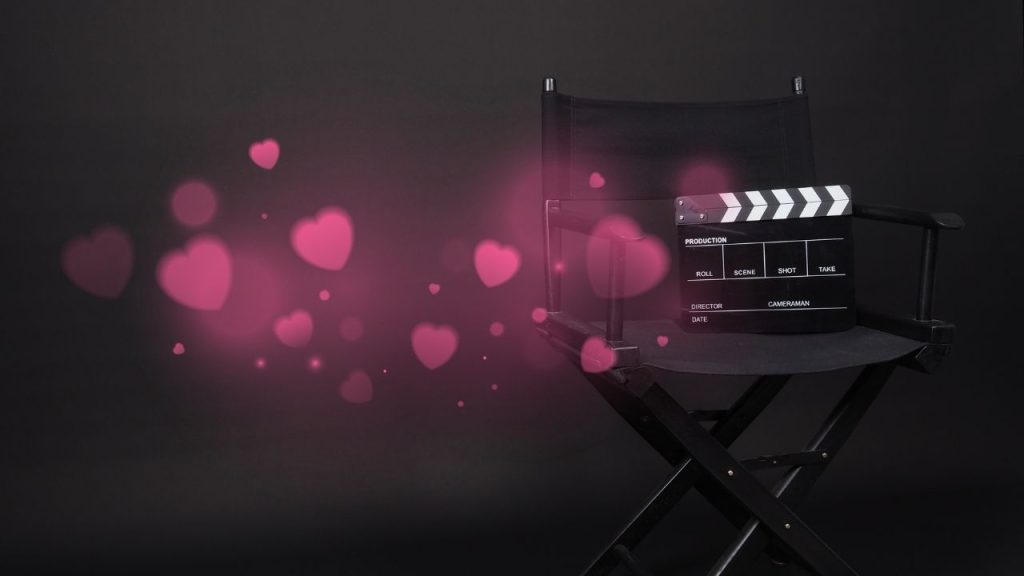 A black and white movie clapperboard sits on a black director's chair against a black background. Overlaid on the image are a group of pink hearts in varying degrees of focus.