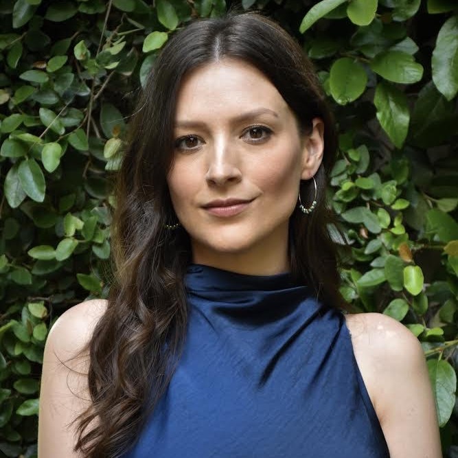 Danica Nava, a light-skinned Chickasaw woman with long, wavy dark hair and a sleeveless blue dress, looks at the viewer while standing in front of a wall of leaves.