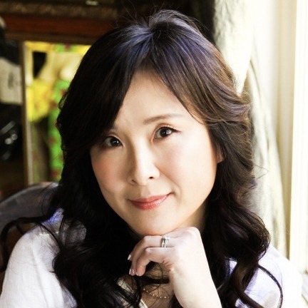 Jayci Lee, a Korean American woman with long, wavy dark hair and a white, long-sleeved button down shirt, looks at the viewer while resting her chin on her hand. A wall and white curtain can be seen behind her.