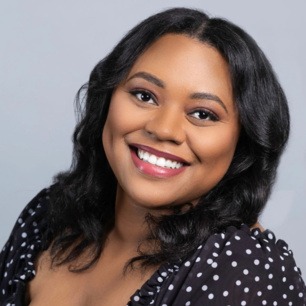 Myah Ariel, a Black woman with wavy shoulder-length dark hair and a black blouse with white polka dots, smiles at the viewer against a gray background.