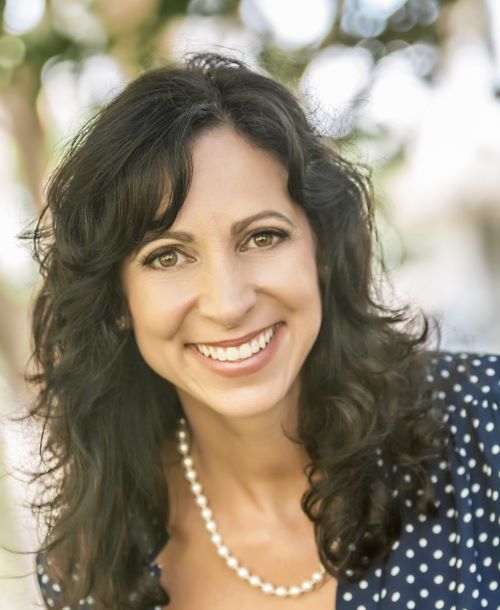 Priscilla Oliveras, a light-skinned Latina woman with long, wavy dark hair and a blue blouse with white polka dots, smiles at the viewer. Trees can be seen out of focus behind her.