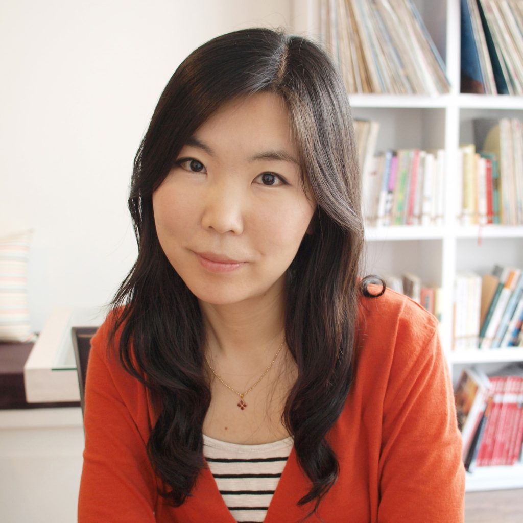 Taeeun Yoo, a smiling light-skinned East Asian woman with long, wavy brown hair and brown eyes, wearing an orange cardigan over a black and white striped blouse, posing in a light-filled room with shelves full of children's books as a background.