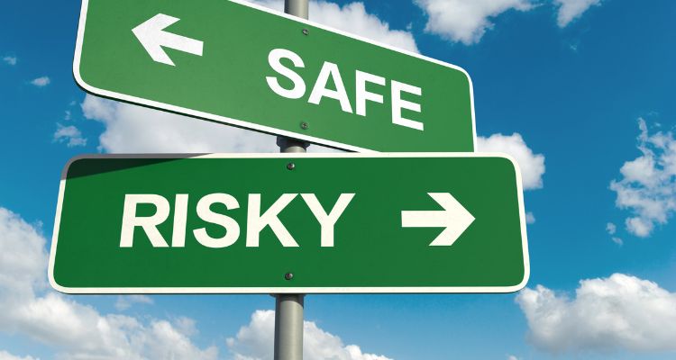 Background is a blue sky with clouds. In the center is a street sign pole with two green signs on it. Pointing to the left is a sign that reads safe, and pointing to the right is one that reads risky.