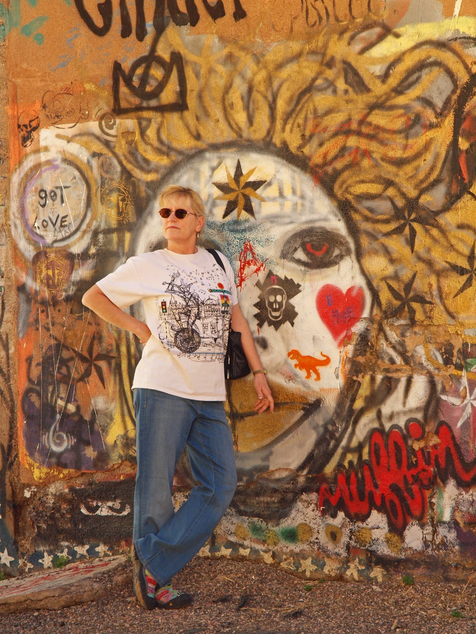 Author Yvonne Navarro, an American woman wearing a white top with illustrations over it, blue jeans, a black shoulder bag, and sunglasses, posing with one hand on her hip, and the other over her bag, looking to the side.