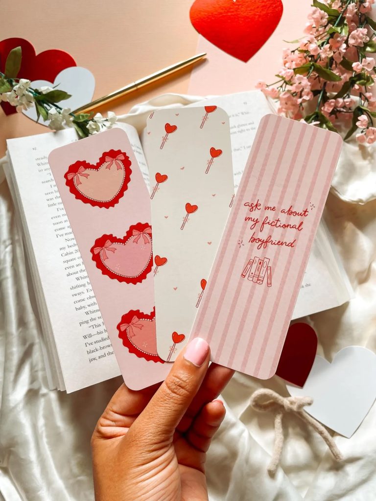 A hand holding a pink bookmark with hearts and bows, a white bookmark with heart-shaped lollipops, and a pink striped bookmark with the words "ask me about my fictional boyfriend" in red cursive letters. The background depicts beige fabric, an open book, and pink and white flowers.