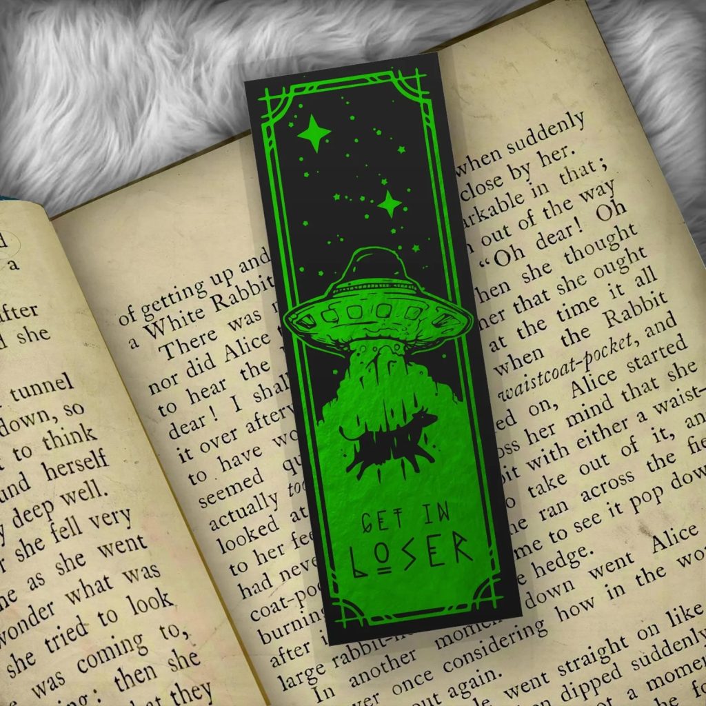 A neon green and black bookmark that depicts a cow being abducted by a UFO, stars, and the words "get in loser" in an otherworldly font. The bookmark is on top of a book page and the book is on top of a grey fuzzy fabric.