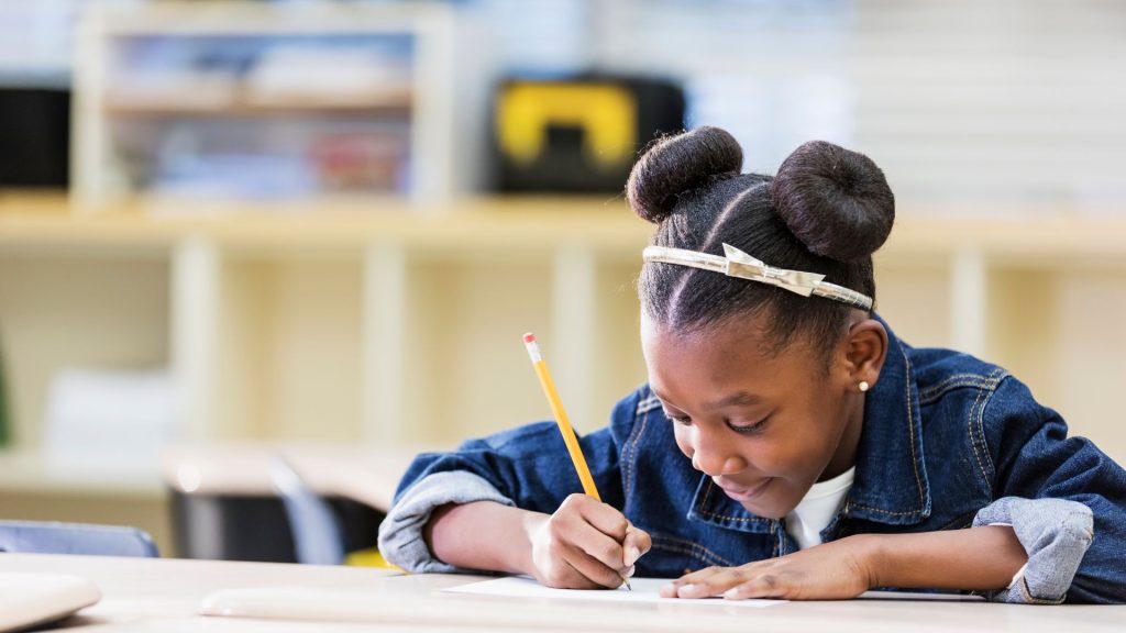 A young Black girl with hair tied in two buns, wearing a denim jacket and bow headband, writes with a #2 pencil on a piece of paper at a desk, cubbies visible behind her.