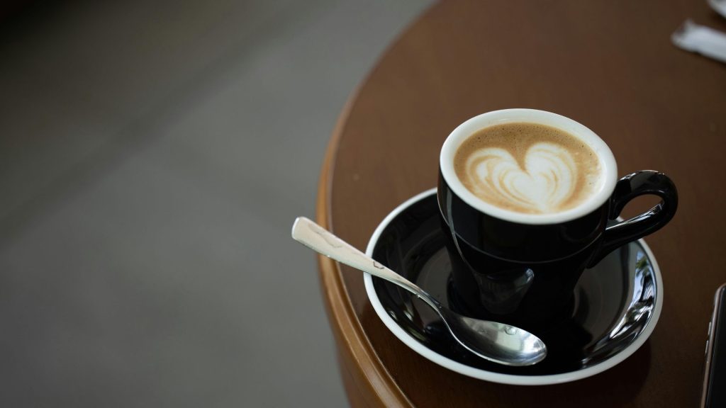 A black mug of coffee on a black saucer with a teaspoon, sitting on a round brown table. A heart is drawn in the foam of the latte.