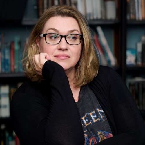Mikki Daughtry, a White woman with short blonde hair and black glasses, wearing a black sweater and graphic T-shirt, looks down to the viewer's right while resting her chin on her hand. She is posing in front of a bookshelf.
