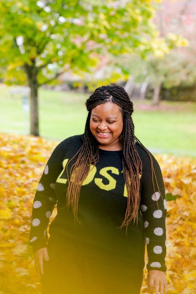 Neena Viel, a Black woman with long braids and a black sweater, smiles while looking down. Behind her is a park, out of focus, with fall leaves and a tree.