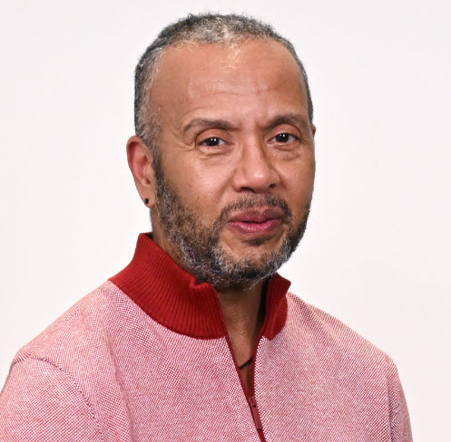 Stacy Nathaniel Jackson, a Black man with short graying hair and graying facial hair, wearing a light red sweater, looks at the viewer. The background is white.
