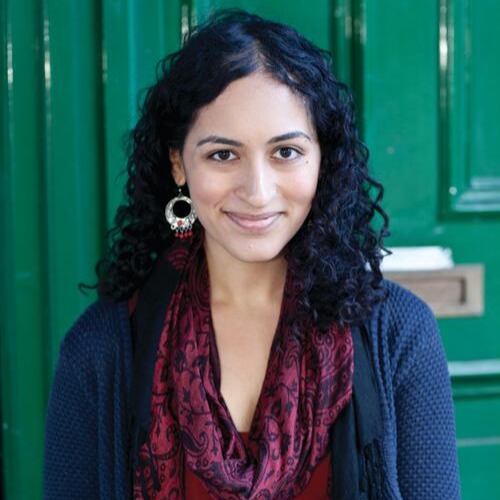 Tasha Suri, a South Asian woman with curly black hair, a blue sweater, and a burgundy scarf, looks at the viewer as she stands in front of a green door.