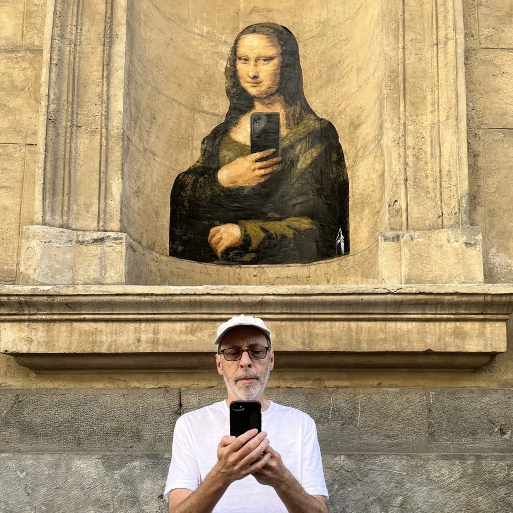 Dan Gutman, a White man with graying stubble, black tinted glasses, a white baseball cap, and a white T-shirt, looks at his phone while standing in front of a wall with an arch carved into it. Painted into the arch is the Mona Lisa holding a smartphone.