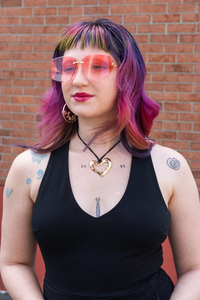 Author Beatrice Lebrun, wearing a black tank top and pink-tinted glasses, with rose gold hoop earrings, and a gold, heart-shaped necklace on a black string, smiling and looking away.