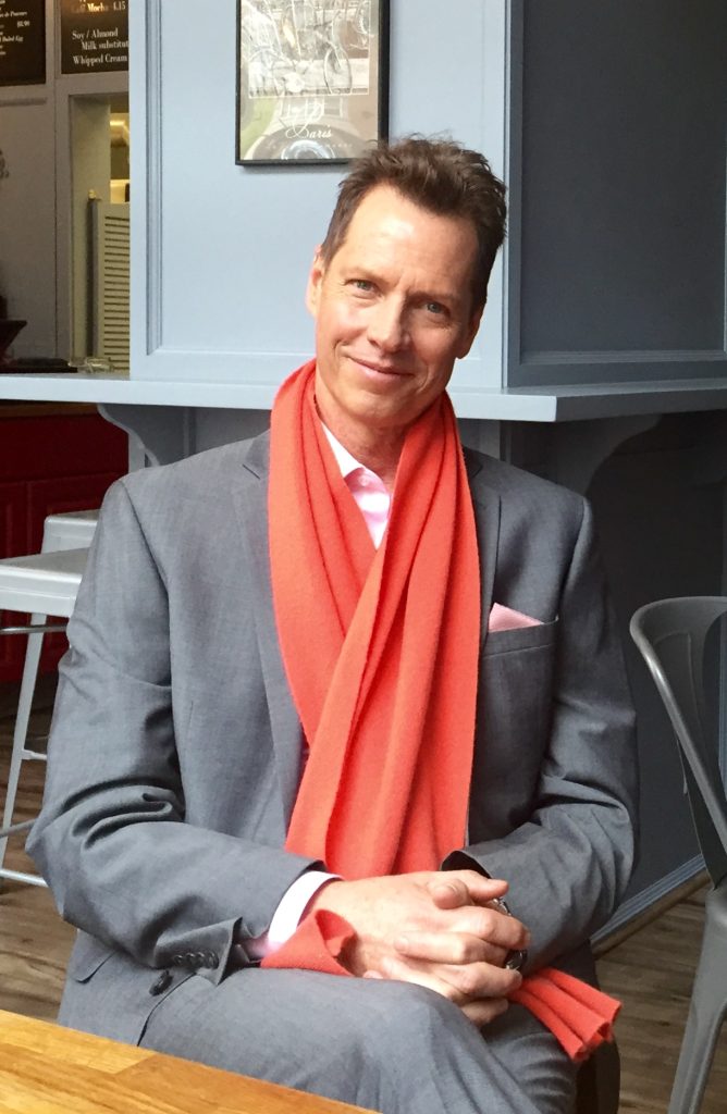 Author Frank Beddor, wearing a grey suit over a pink shirt, with a vivid, orange scarf wrapped around his neck, hands folded, smiling and looking ahead.