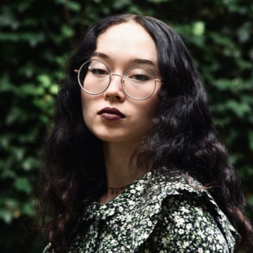 Author Zoe Hana Mikuta, a Korean woman, wearing a dark toned, floral designed top, with a black choker necklace, and round shaped glasses, looking ahead.