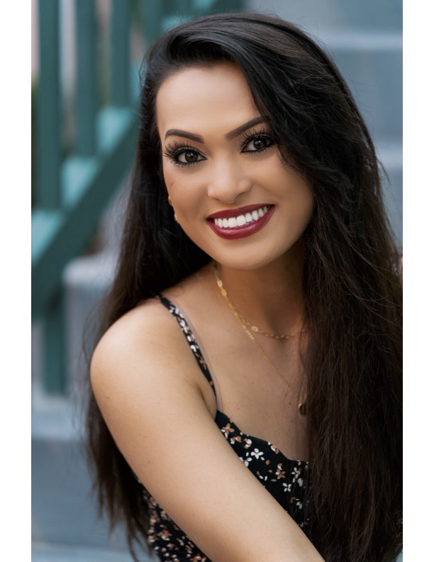 Headshot of the romantasy cosplayer Jane. It is a close up of her head and shoulders. She has long black hair and a large smile. There is a blurred background of steps.